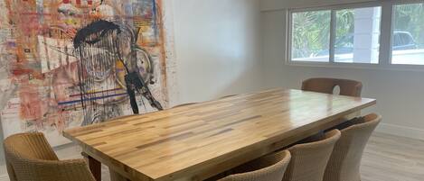 Un comedor con mesa de madera y sillas de mimbre, un cuadro abstracto grande en la pared, y una ventana con vistas al exterior.