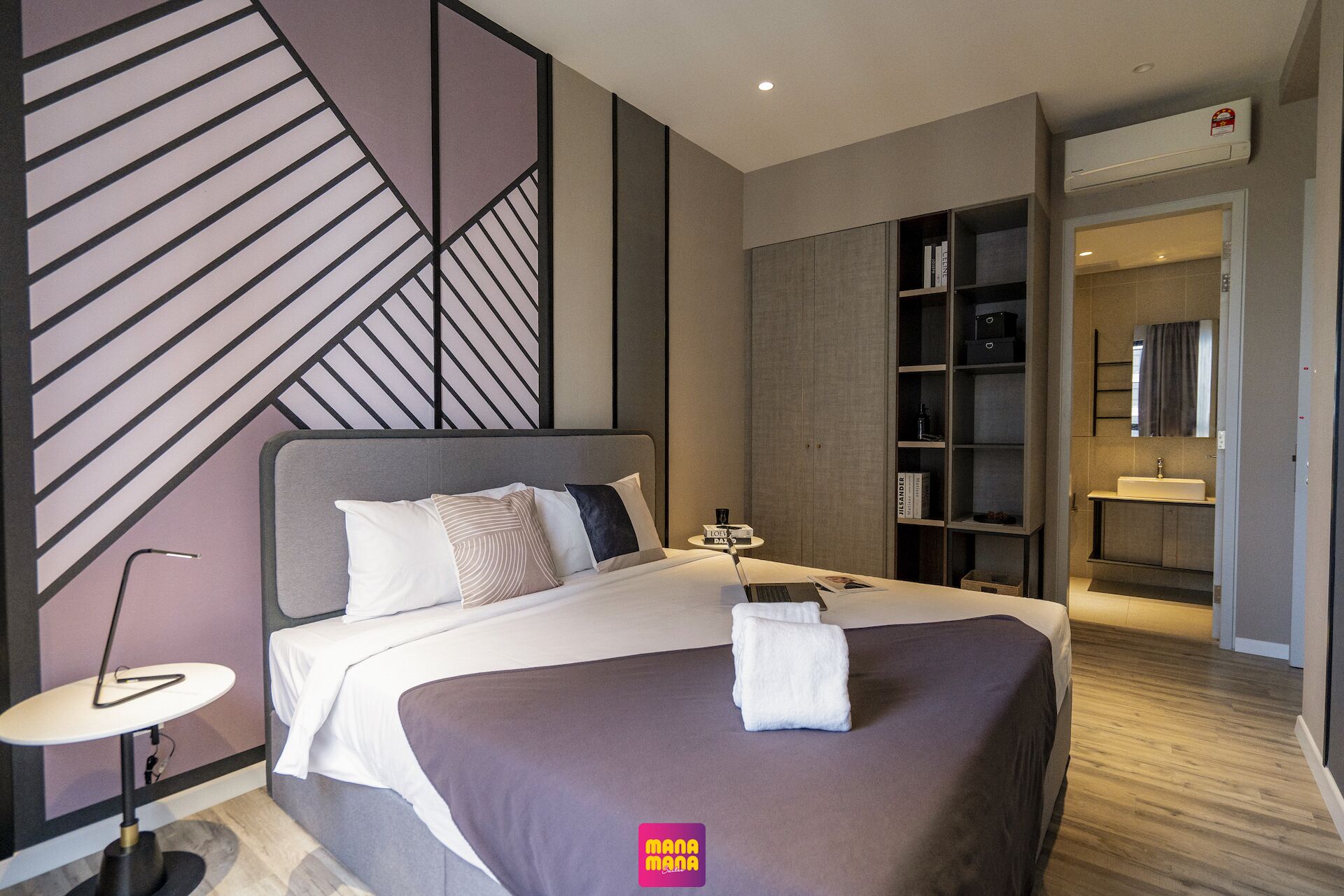 A modern hotel room with a large bed, a bedside table, a small round table, a bookshelf, and a bathroom visible through an open door.