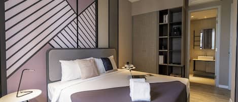 A modern hotel room with a large bed, a bedside table, a small round table, a bookshelf, and a bathroom visible through an open door.