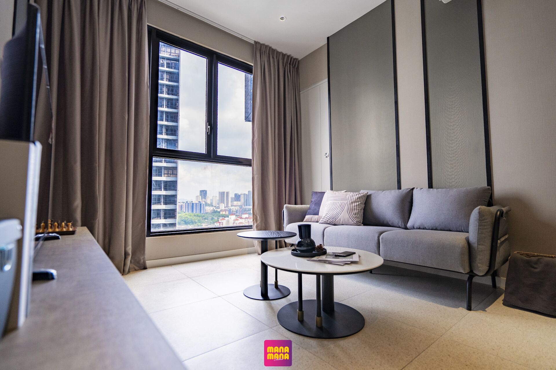 Un salon moderne avec un canapé gris, une petite table ronde et une vue sur le paysage urbain à travers de grandes fenêtres.
