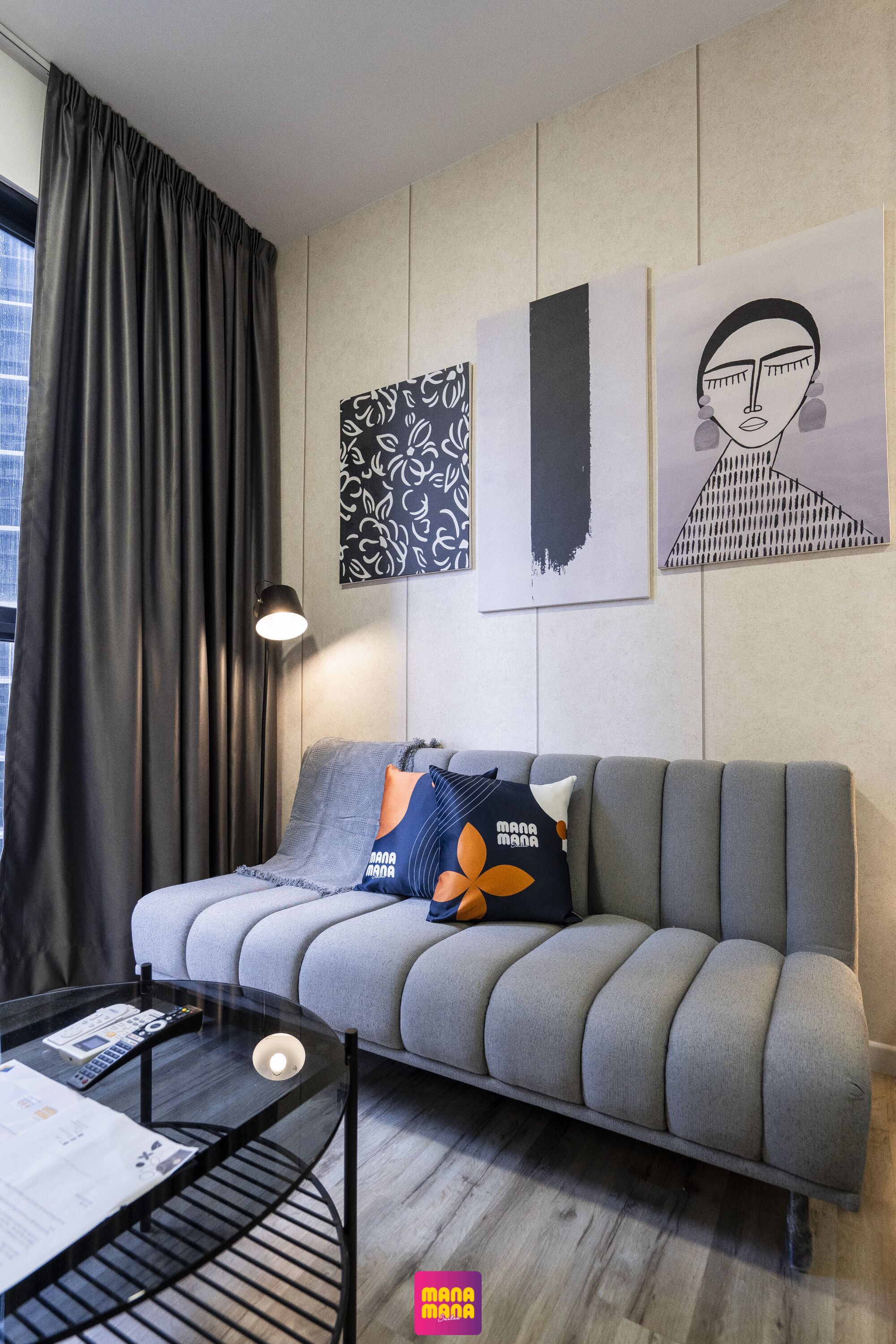 Un saló modern amb un sofà gris, una taula de centre de vidre i una làmpada. Hi ha elements decoratius abstractes a la paret i una finestra amb cortines.