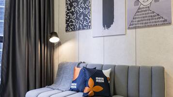 Un saló modern amb un sofà gris, una taula de centre de vidre i una làmpada. Hi ha elements decoratius abstractes a la paret i una finestra amb cortines.