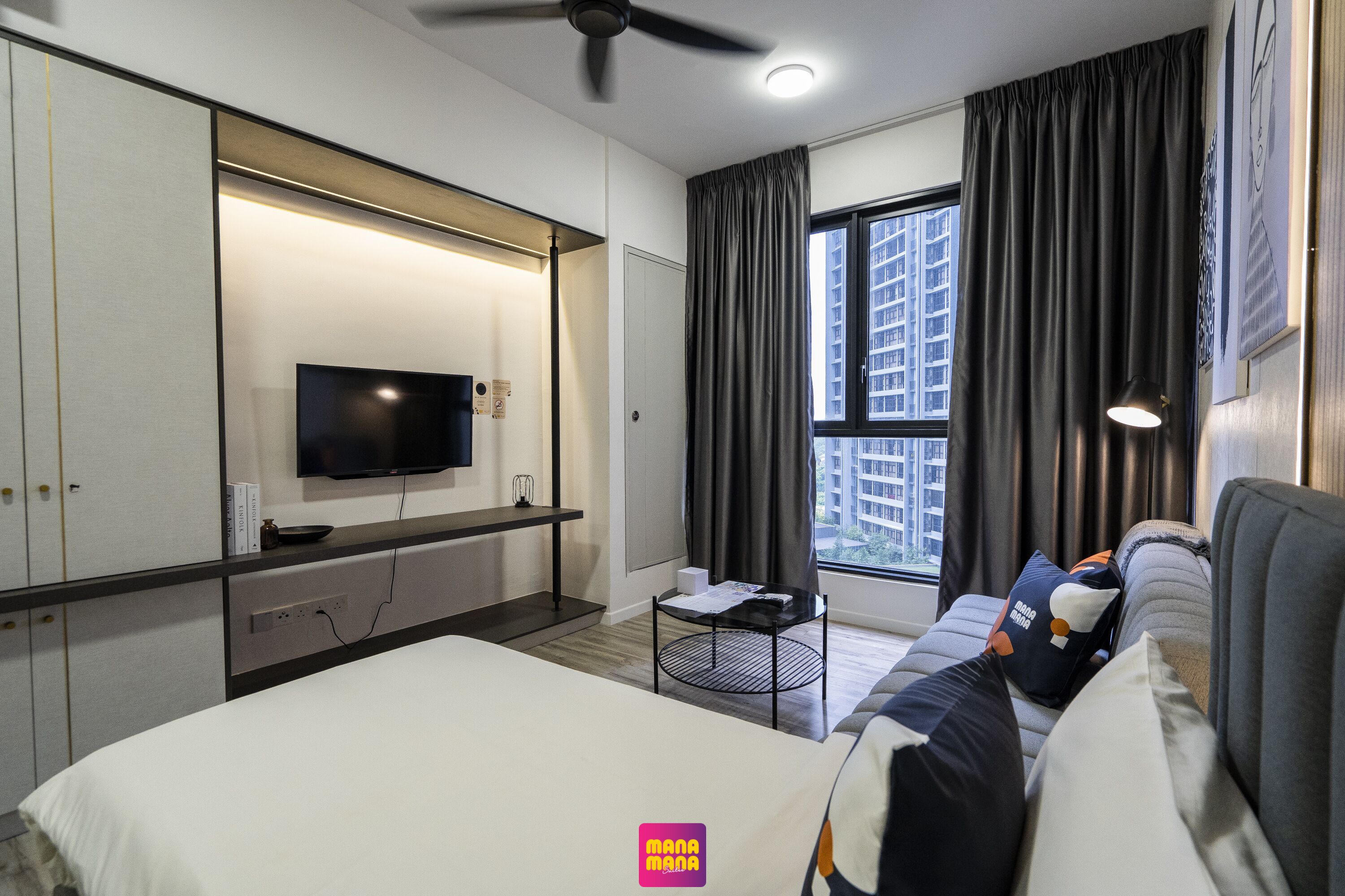 A hotel room with a bed, a TV mounted on the wall, a window with curtains, a sofa with pillows, and a ceiling fan.