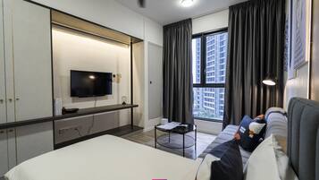 A hotel room with a bed, a TV mounted on the wall, a window with curtains, a sofa with pillows, and a ceiling fan.