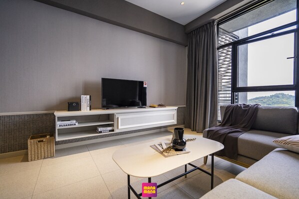 A modern living room with a flat-screen TV, a coffee table, a sofa, and a large window offering a view of the outdoors.