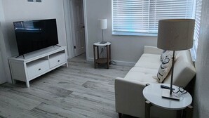 A living room with a flat-screen TV on a white stand, a beige sofa, a small round table with a lamp, and a ceiling fan.