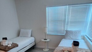 A room with two beds, a ceiling fan, a small table, and a window with blinds.
