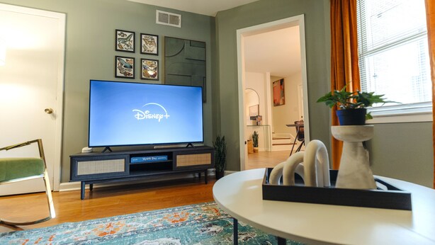 Un salon avec un téléviseur à écran plat diffusant Disney+, un canapé, une table basse et un tapis à motifs.