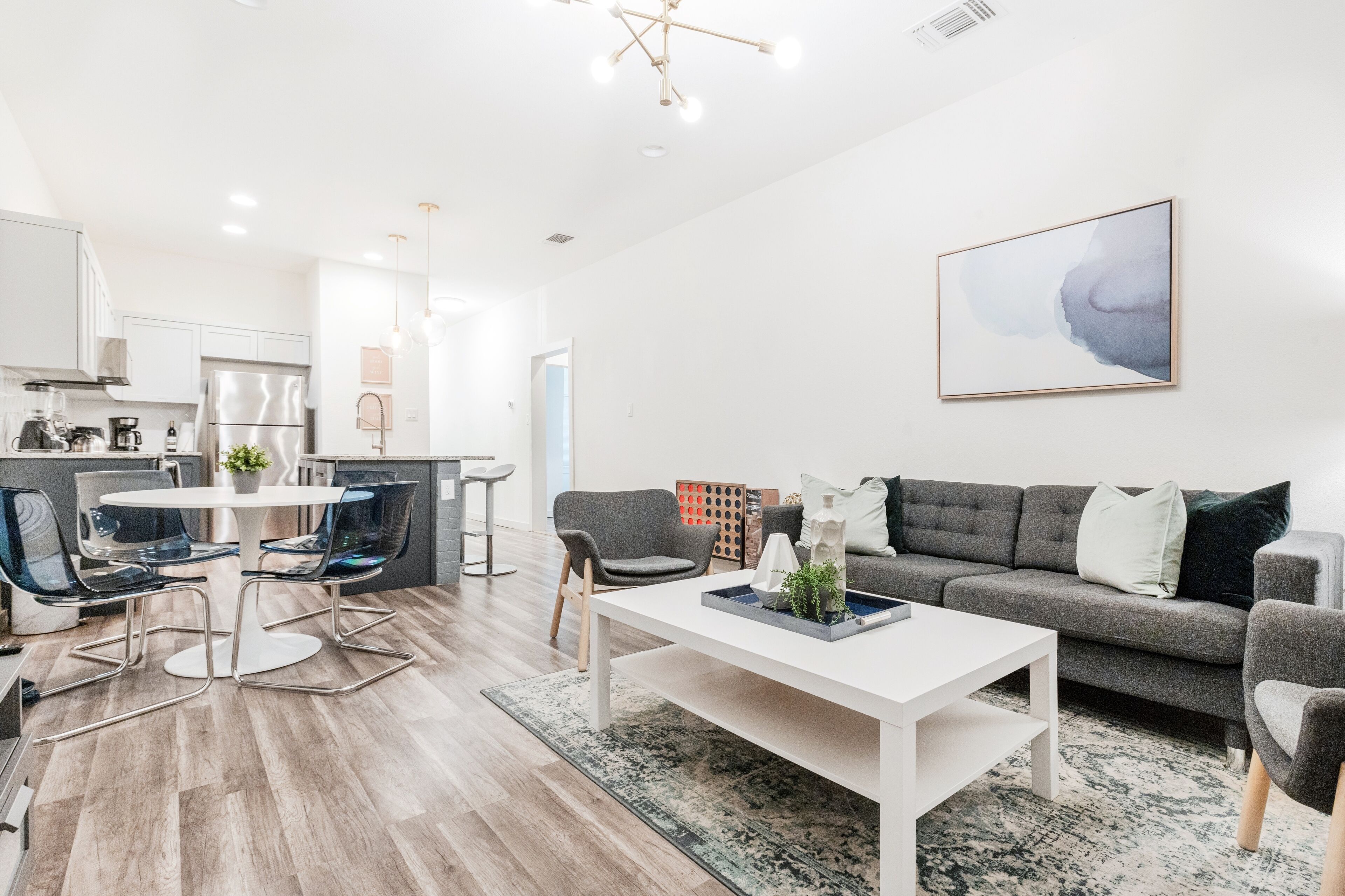 Un salon moderne avec une table basse blanche, un canapé gris et des chaises bleues. On aperçoit une cuisine en arrière-plan, avec des meubles blancs et des appareils électroménagers en acier inoxydable.
