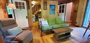 Une pièce chaleureuse avec un plancher en bois, un canapé vert, un fauteuil en bois et une petite table. Un escalier mène à un autre niveau, et une porte est dotée de panneaux de verre.