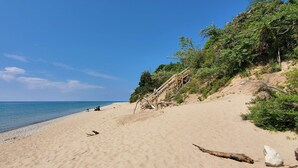 Une plage avec un escalier en bois menant au sommet d’une pente de sable, un ciel bleu clair et une mer calme.