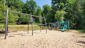 A playground with swings and a slide, surrounded by trees and a sandy ground.
