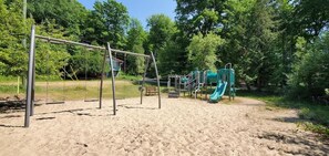 A playground with swings and a slide, surrounded by trees and a sandy ground.