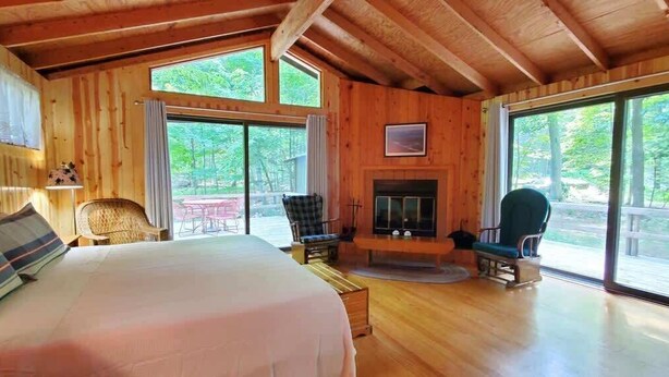 A wooden cabin with a large bed, two chairs, a fireplace, and large windows.