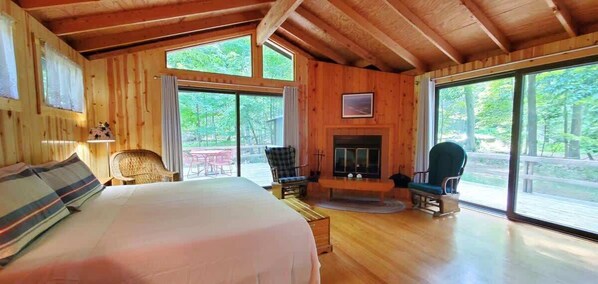 A wooden cabin with a large bed, two chairs, a fireplace, and large windows.