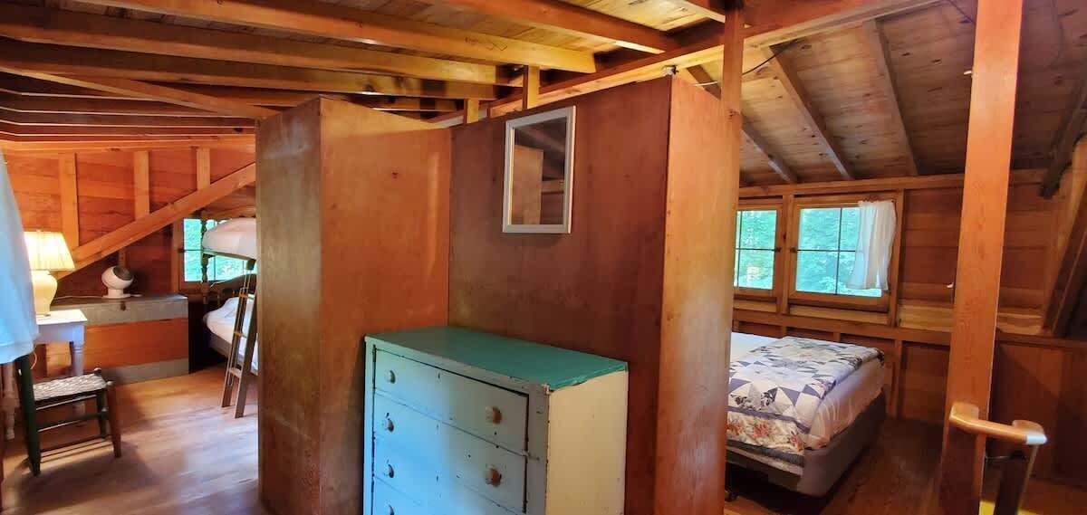 A rustic cabin interior with a wooden ceiling, a bed with a floral bedspread, a dresser, and a window with curtains.