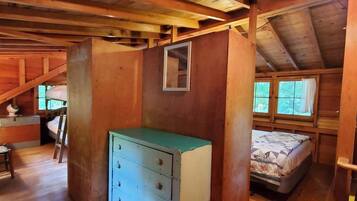 A rustic cabin interior with a wooden ceiling, a bed with a floral bedspread, a dresser, and a window with curtains.