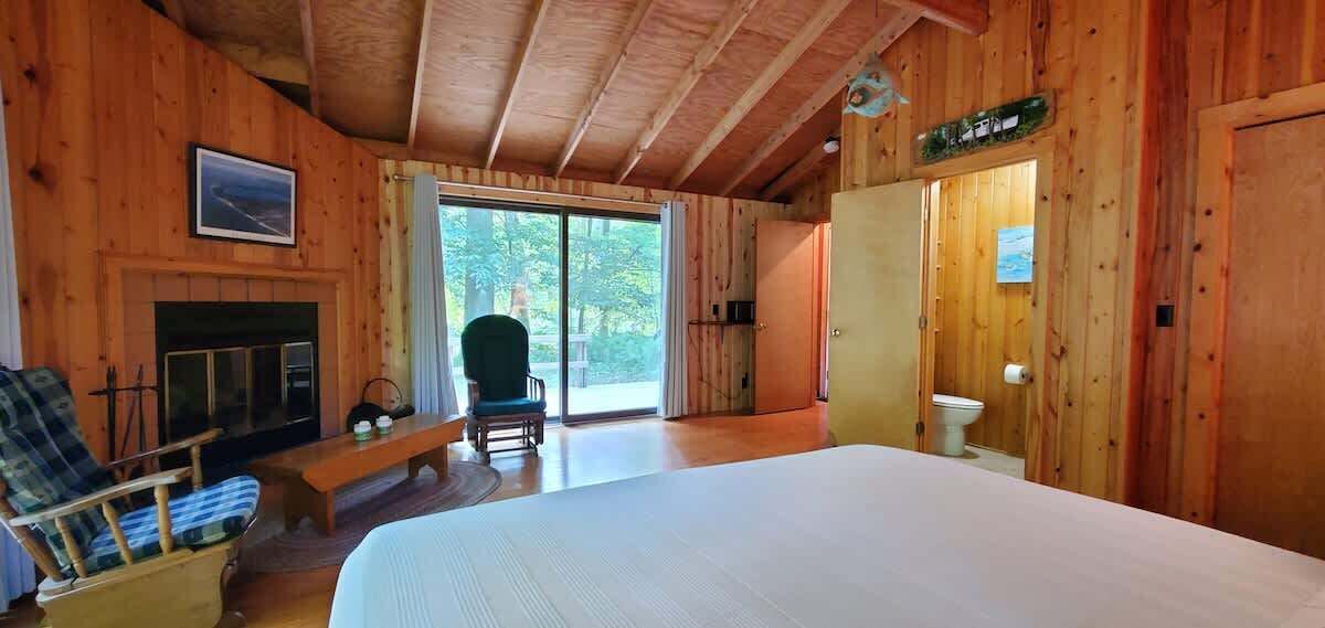 A wooden cabin with a large bed, a fireplace, a bench, and a view of the outdoors.