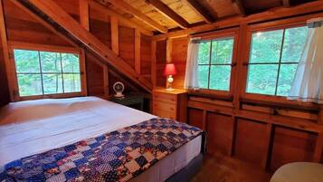 A cozy wooden cabin bedroom with a bed, a nightstand, a lamp, and two windows with curtains.