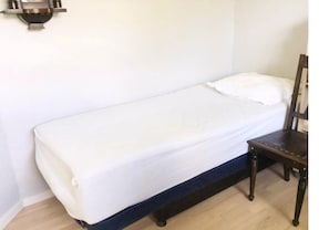 A single bed with a white mattress and pillow, a wooden chair, and a wall-mounted light fixture.