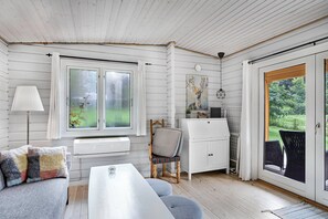 A room with a white sofa, a dining table, a chair, a lamp, a window, and a view of greenery outside.