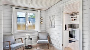 Habitación con dos sillas, una mesita, una ventana con cortinas, un reloj de pared, un cuadro y vistas a una zona de cocina.
