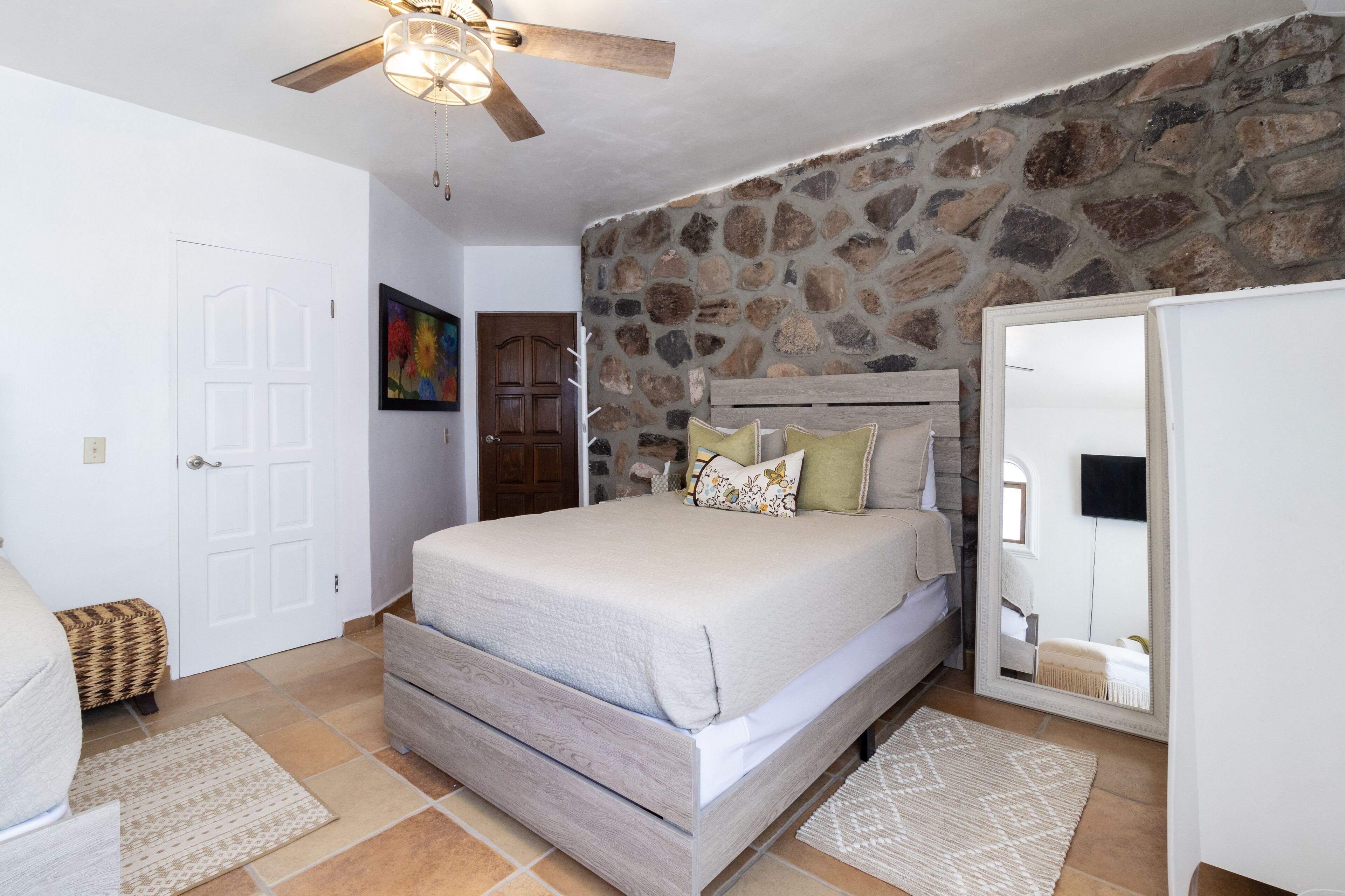A bedroom with a stone wall, a bed with a wooden headboard, a ceiling fan, and a large mirror.