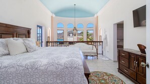 A bedroom with a large bed, a chandelier, a view through arched windows, and a balcony.