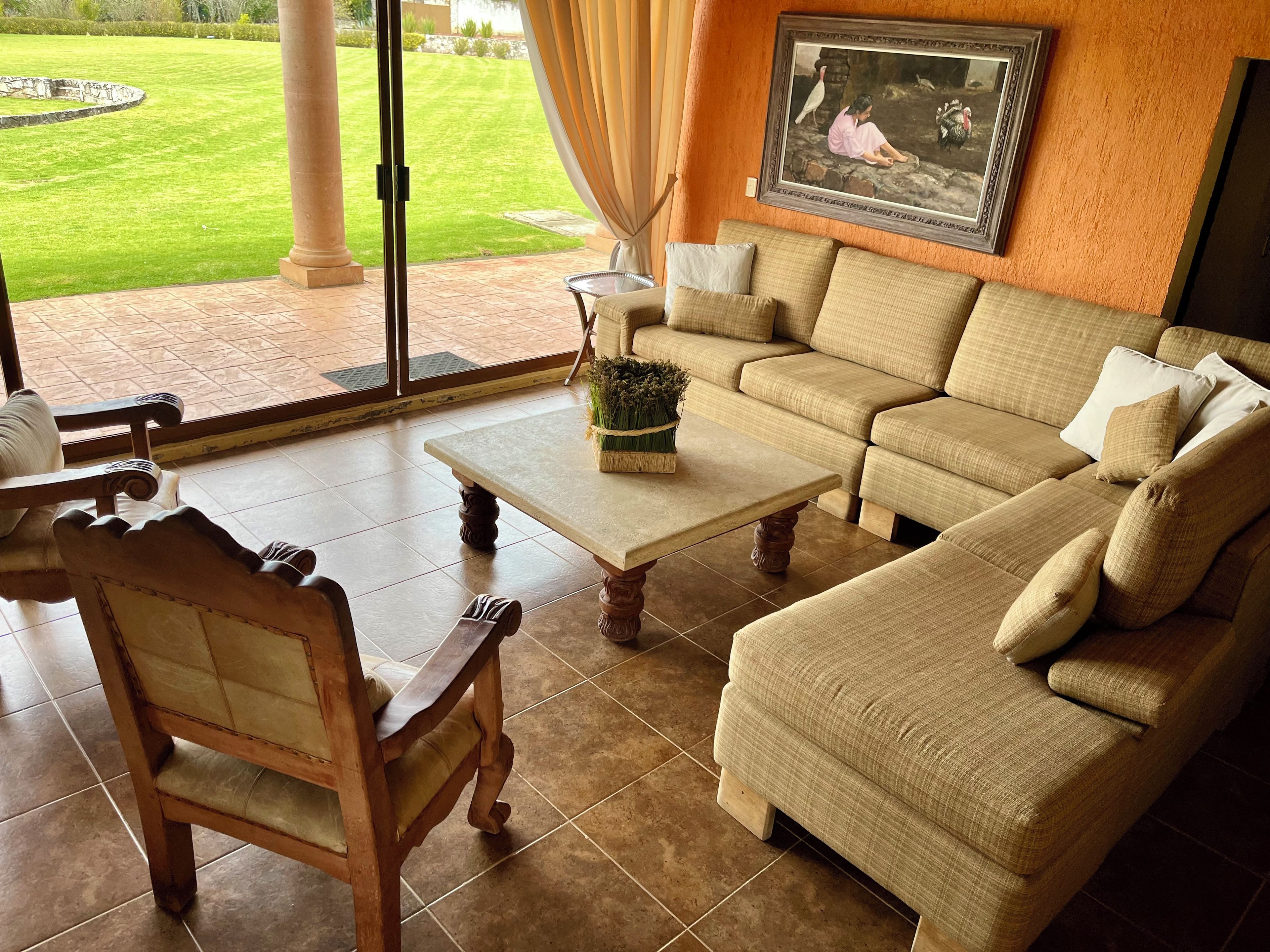 A living room with a sofa, armchair, and coffee table, a painting on the wall, and a view of a green lawn through a glass door.