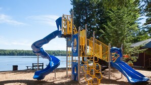A playground with two blue slides and yellow safety railings, set against a backdrop of trees and a lake.