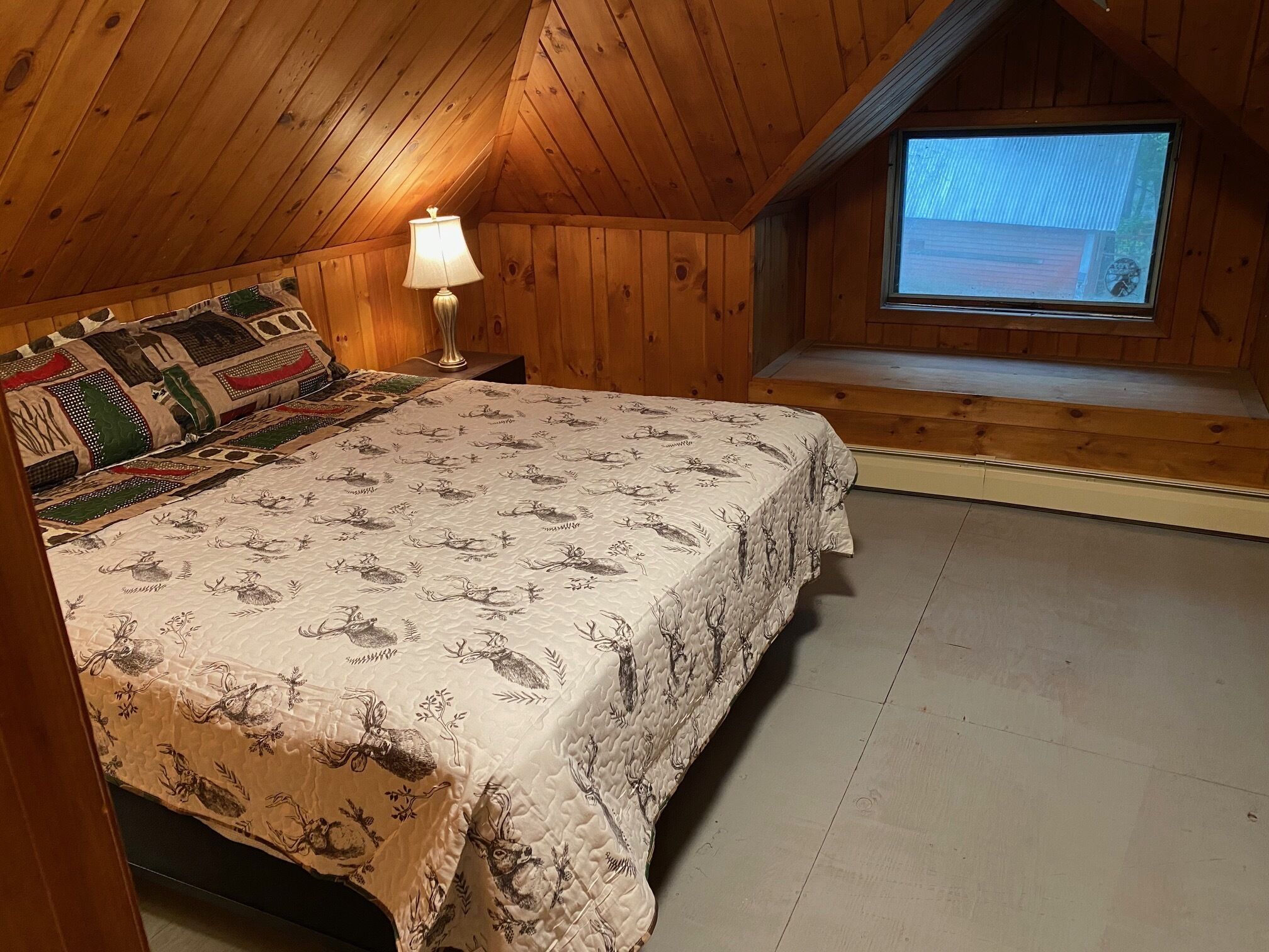A cozy wooden room with a bed, a nightstand, a lamp, and a window.