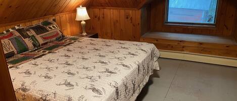 A cozy wooden room with a bed, a nightstand, a lamp, and a window.