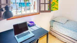 Une pièce avec un ordinateur portable sur un bureau, un lit recouvert d’une couverture à motifs, une fenêtre donnant sur la plage et un tableau coloré accroché au mur.
