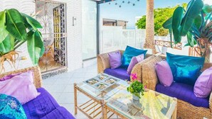Un espace extérieur accueillant, aménagé avec des meubles en osier, des coussins violets et une table à plateau en verre.