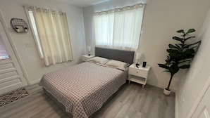 Un dormitorio con una cama, mesitas de noche, una planta en maceta y una ventana con cortinas.