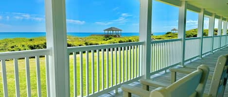 A covered porch with white railings and wooden benches, overlooking a grassy area with a gazebo and the ocean.