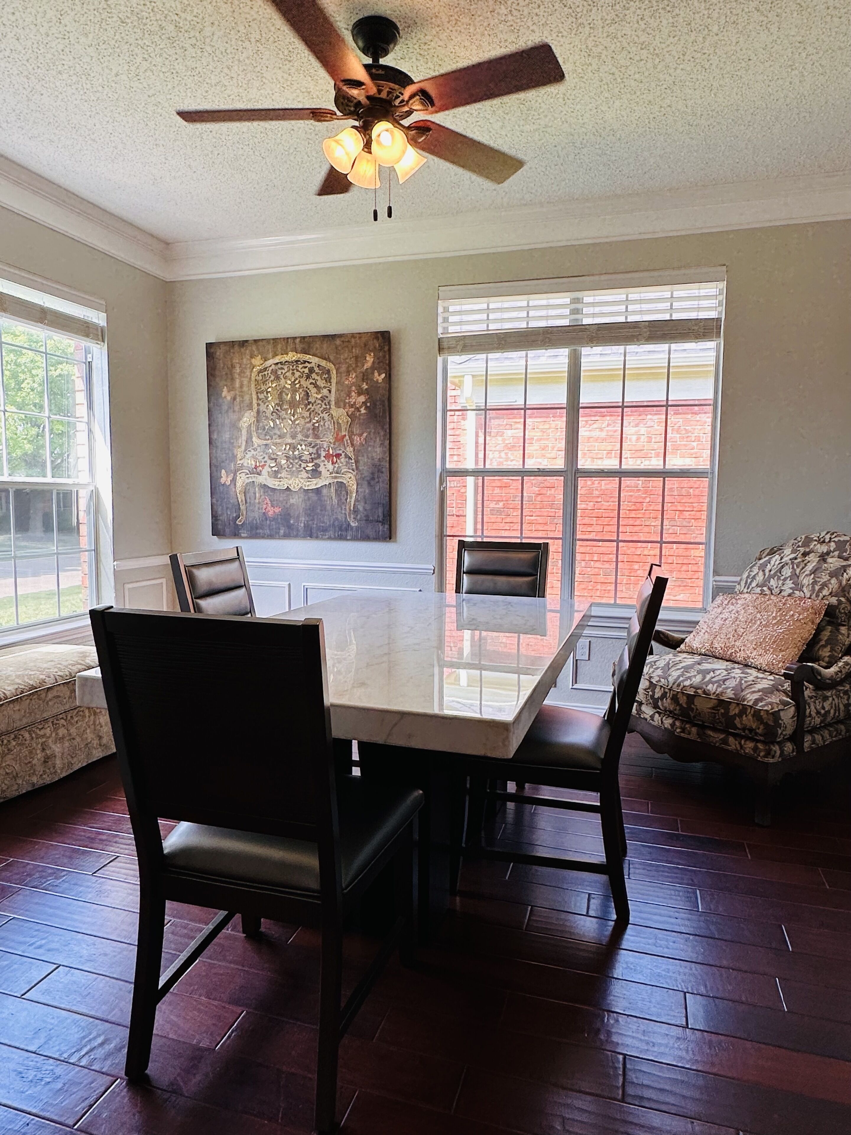 A dining area with a large table, chairs, a ceiling fan, and a window with a view of a brick building.