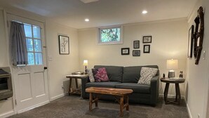 A cozy living room with a grey sofa, a wooden coffee table, and a small side table with a lamp. There are framed pictures on the wall and a window with curtains. - Private Apartment on Beautiful Grounds, Walking Distance to Landrum SC (Landrum)