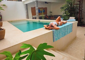 A pool area with a person lying on a ledge, a sofa, and a staircase with potted plants.