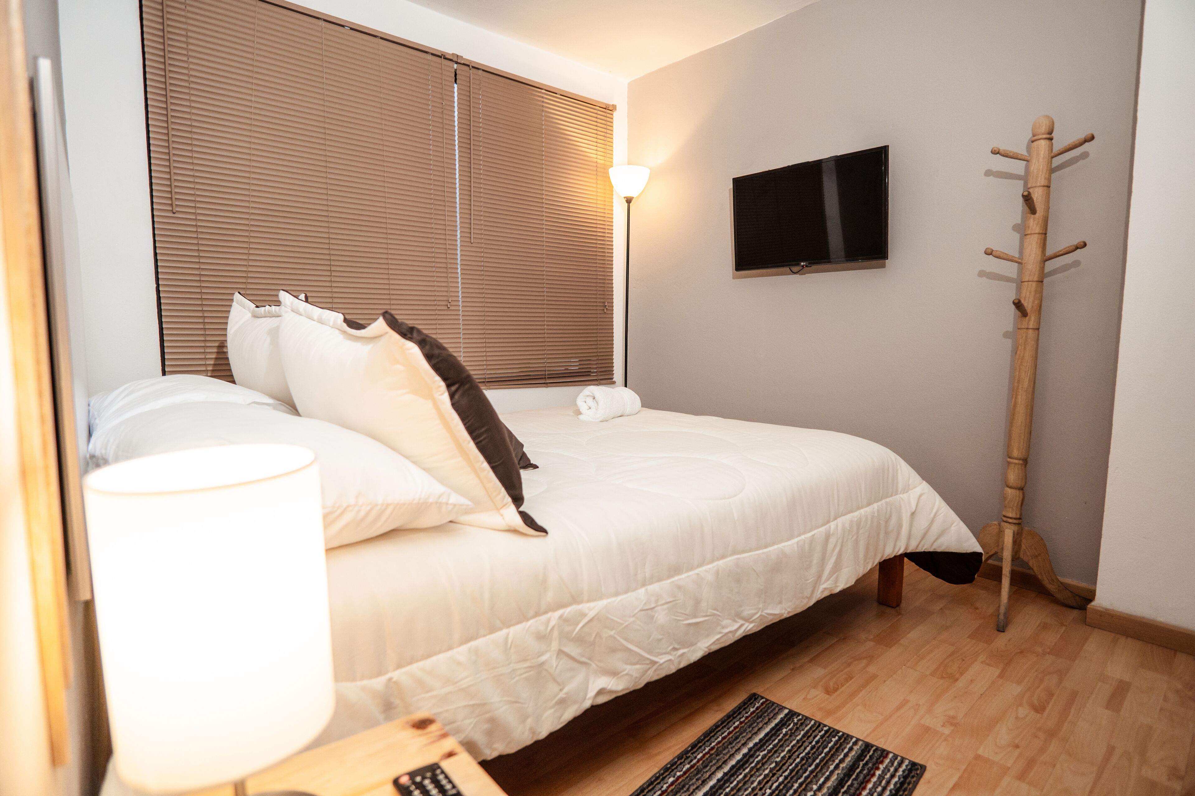 A neatly arranged bedroom with a bed, a bedside lamp, a television mounted on the wall, and a wooden coat stand.
