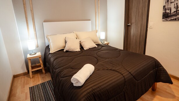 A neatly made bed with a brown bedspread and multiple pillows, a wooden nightstand with a lamp, and a wooden floor. - Primitivo Usaquén dn (Bogotá)