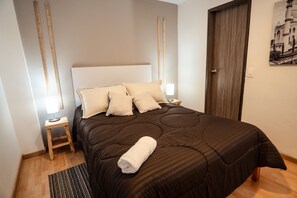 A neatly made bed with a brown bedspread and multiple pillows, a wooden nightstand with a lamp, and a wooden floor.