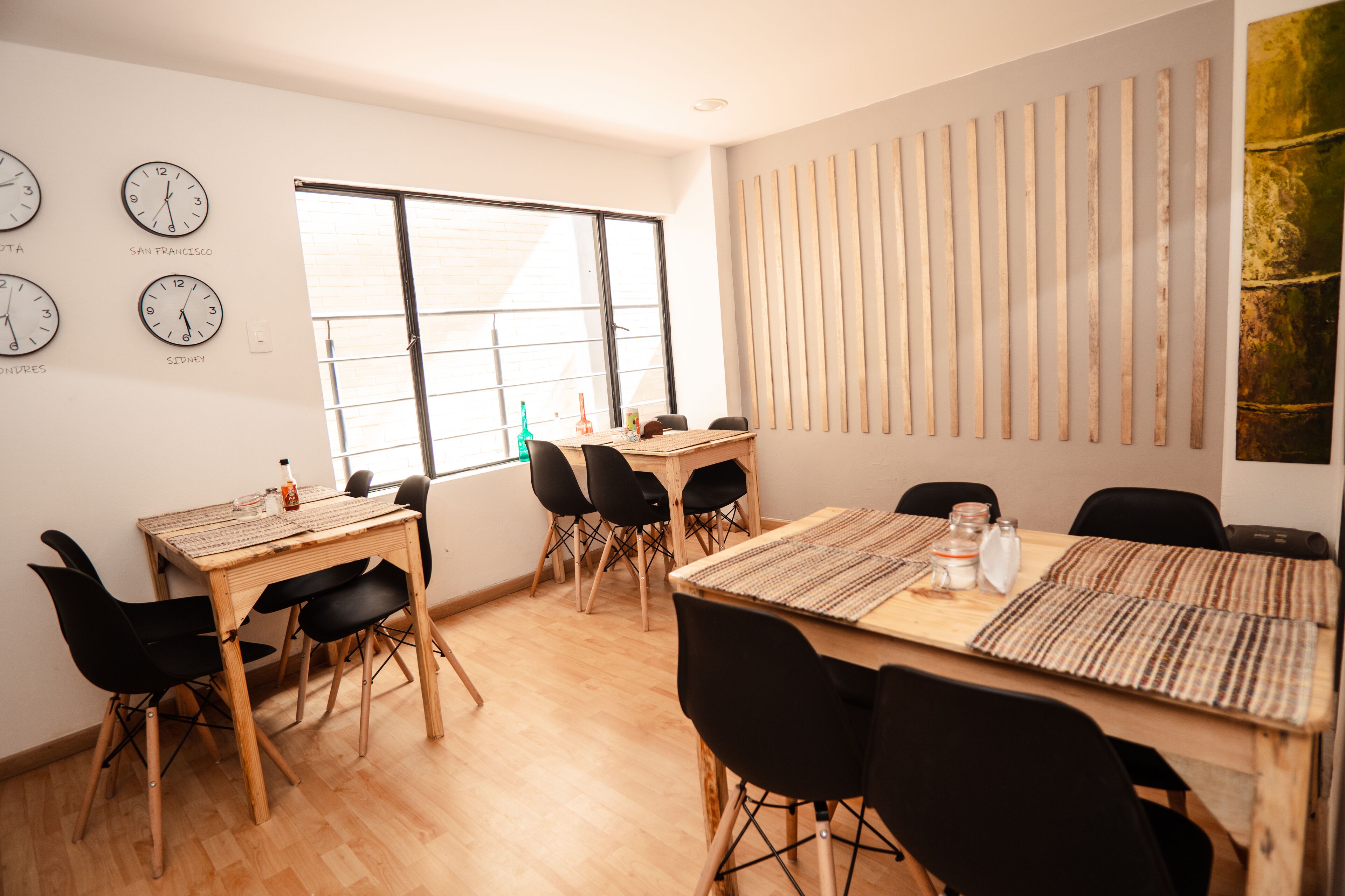 A modern dining area with wooden tables and black chairs, large windows, and wall clocks.