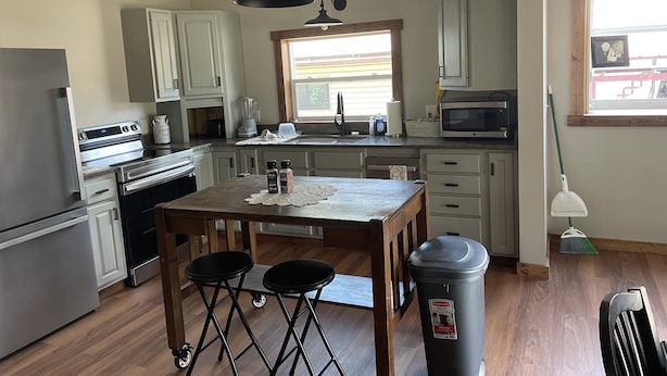 Una cocina con armarios blancos, una mesa de madera, dos taburetes, un cubo de basura y una ventana con persianas.