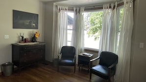 Una habitación con suelo de madera, dos sillas negras, una mesita, un armario de madera oscura, un cuadro en la pared y cortinas blancas.