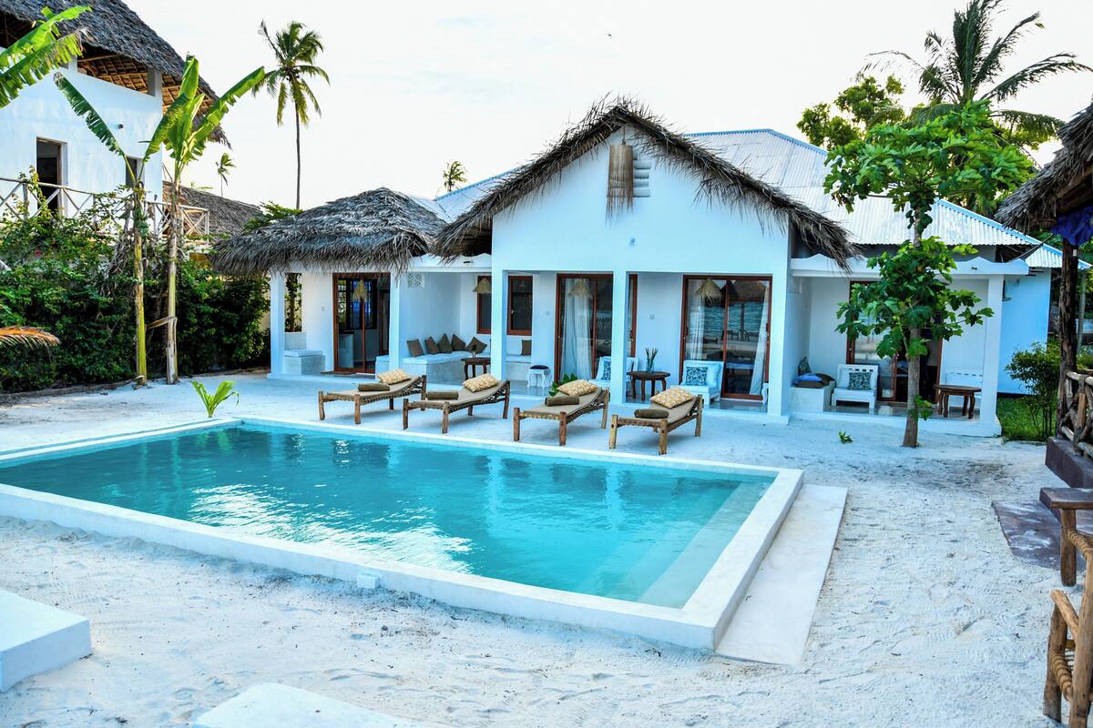 A poolside area with lounge chairs, a swimming pool, and a thatched-roof building.