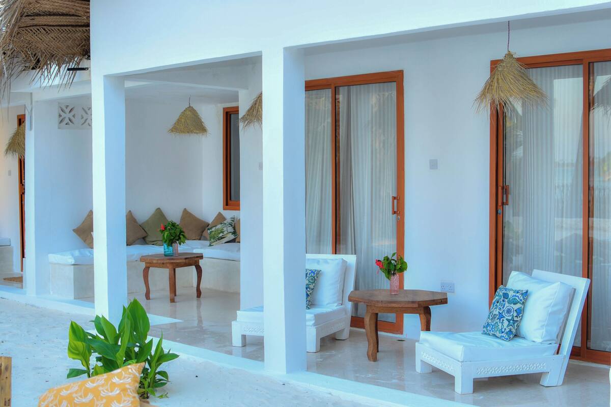 A white outdoor seating area with cushioned chairs, a wooden table, and a potted plant.