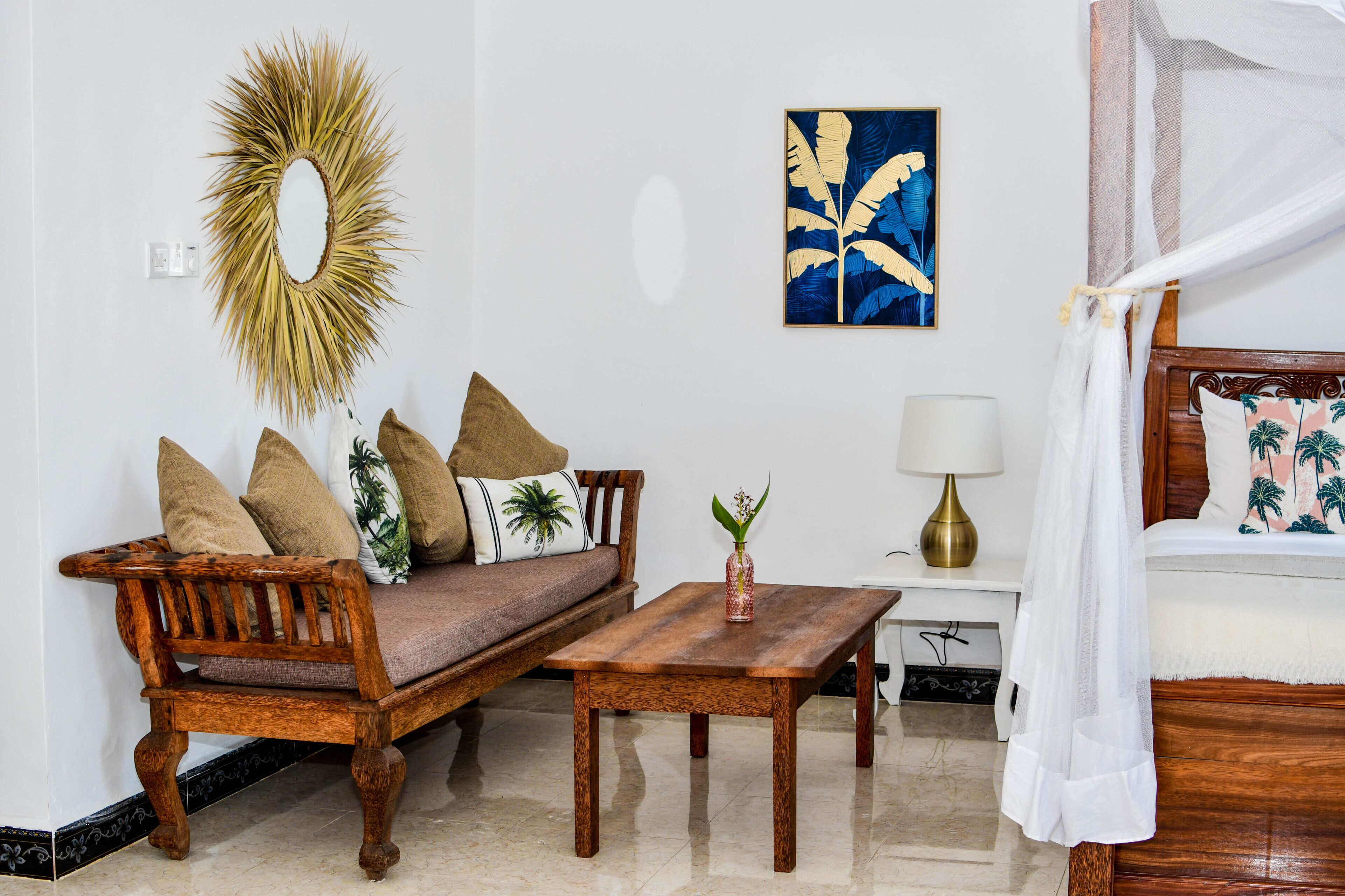 Un dormitorio con una cama y un sofá de madera, una mesita, una lámpara, un espejo decorativo y un cuadro en la pared.