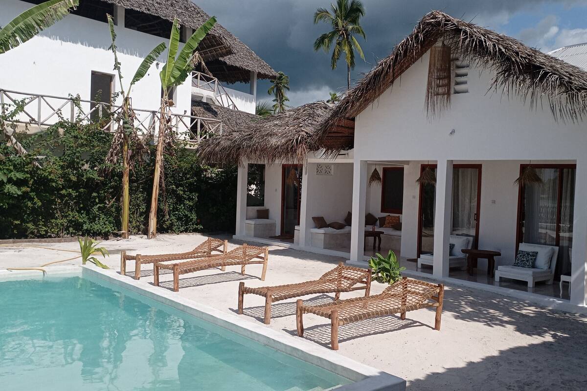 A beachfront property with a pool, lounge chairs, and a thatched-roof building.