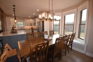 A spacious kitchen with a large island, wooden dining table, and chairs. There are multiple windows providing natural light and views of the outdoors.